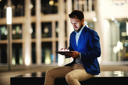 Trendy formal man in suit sitting against illuminated building in night time and browsing tabletの写真素材