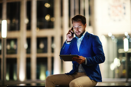 Adult stylish man watching tablet while speaking on phone doing business and sitting outdoors in night lightsの写真素材