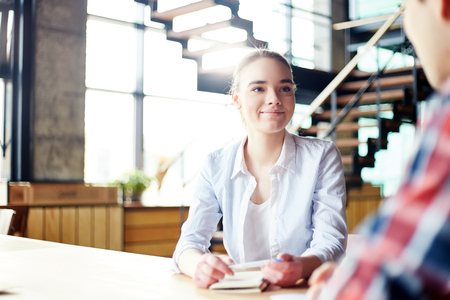 Young woman in casual clothing sitting at table and smiling to anonymous man during meeting in cafeの写真素材
