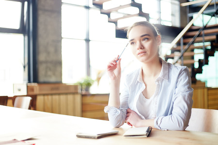 Young woman in white blouse contemplating on assignment while sitting over notepad at desk with pen in her handの写真素材