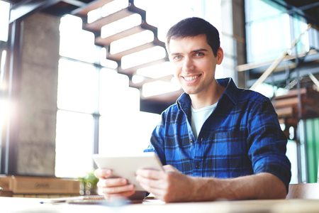 Handsome young man in checkered blue shirt holding touchpad computer and smiling at cameraの写真素材