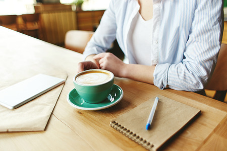 Faceless shot of woman in shirt sitting at table with coffee cup and notebooksの写真素材