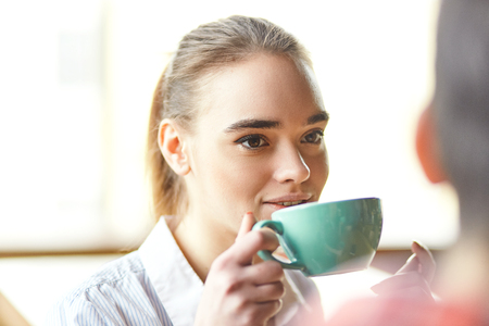 Young blond girl taking sip of coffee while talking to crop anonymous partner in daylightの写真素材