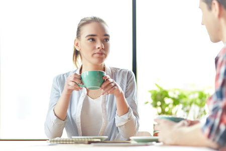 Young casual girl sitting with guy and drinking coffee together with studies on deskの写真素材