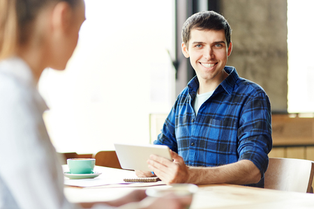 Casual cheerful man in shirt holding tablet and notepad sitting at desk with girl and smiling at camera having coffeeの写真素材