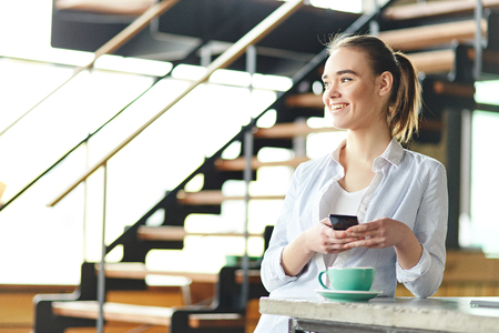 Pretty young girl standing with cup of coffee on counter and holding smartphone while laughing awayの写真素材