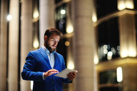 Trendy bearded man browsing tablet standing on street in lights of buildings during night timeの写真素材