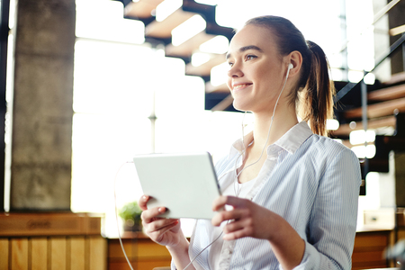 Pretty casual girl holding touchpad and listening to music with earphones smiling away in daylightの写真素材