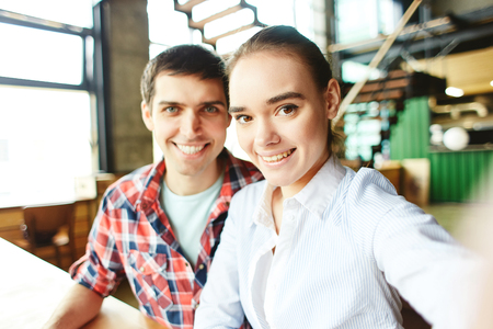 Pretty girl and handsome man posing at camera taking selfie and smiling happily sitting at tableの写真素材
