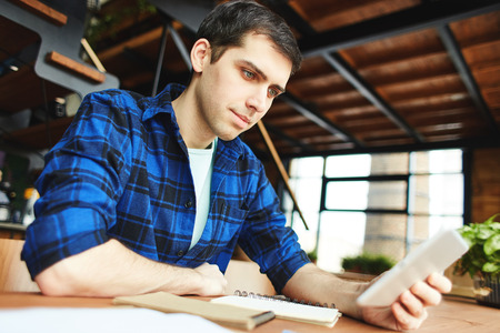 Young man in blue shirt watching tablet sitting at table with notepad and doing studiesの写真素材