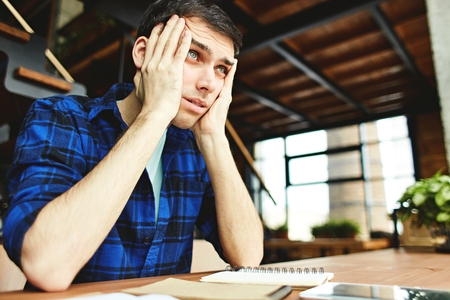 Casual young man leaning on hands sitting with studies at desk unable to find solution and looking awayの写真素材