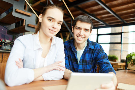 Millennial man and woman sitting at desk with tablet computer and smiling at camera confidentlyの写真素材