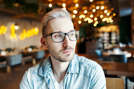 Portrait of handsome young man in eyeglasses sitting with thoughtful facial expression in illuminated cafe aloneの写真素材