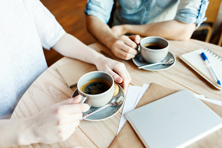 Hands of unrecognizable man and woman having coffee during business meeting in cafe, tablet computer, notepad and papers on table nearbyの写真素材