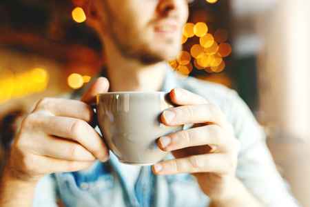 Close-up of unrecognizable man holding cup of coffee or tea in his hands, bokeh lights around himの写真素材