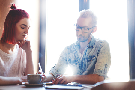 Two friends sitting at cafe table in daylight. Young man in eyeglasses explaining something to his girlfriend, helping her to do homework or sharing his business ideas over cup of coffeeの写真素材