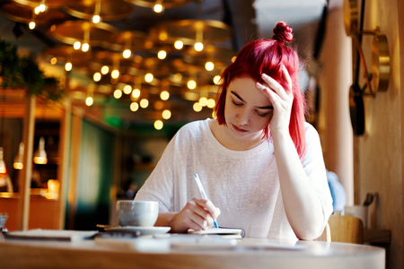 Concentrated young woman writing in notebook while sitting at table in cafe. Female college student with red hair doing her homeworkの写真素材