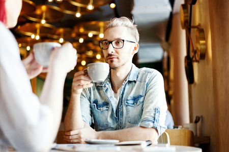 Young man in eyeglasses sitting at table in cafe face-to-face with his unrecognizable girlfriend. Couple drinking coffee and talking.の写真素材