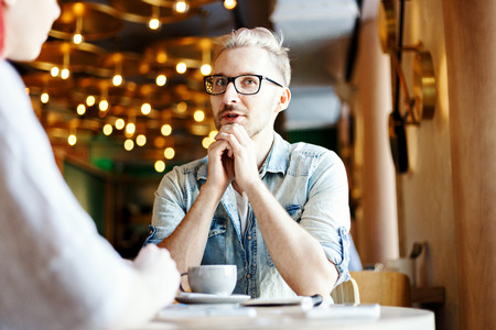 Young man talking to his unrecognizable female friend in cafe. Guy telling story or giving interview over cup of coffee.の写真素材