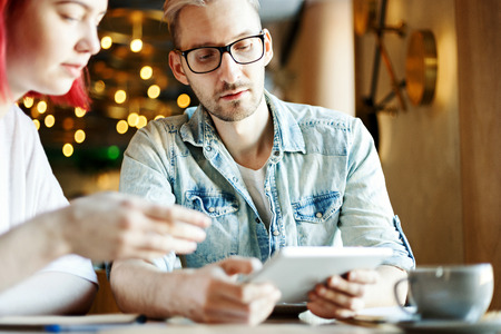 Portrait of man showing something on tablet computer to his female colleague. Friends sitting at table in cafe and using tablet pc together, focus on manの写真素材