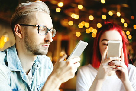 Young couple sitting in cafe together, man and woman focused on their cell phones during date: texting, browsing Internet or using appsの写真素材