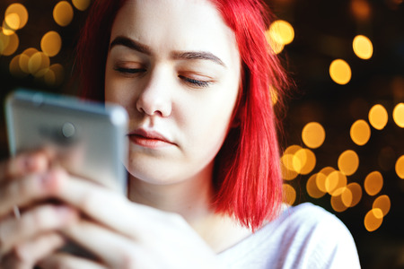 Close-up portrait of girl with red hair text messaging on her smartphone on the background of bokeh...