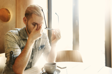 Exhausted young man taking off eyeglasses and rubbing his tired eyes while sitting at cafe table in sunlightの写真素材