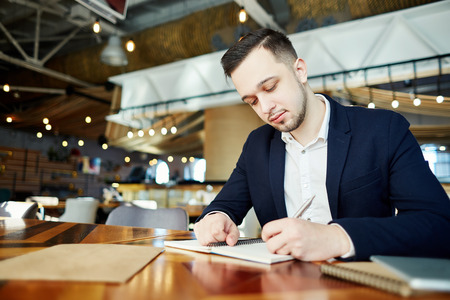 Restaurant manager in shirt and jacket sitting at table and making notes in his personal organizerの写真素材