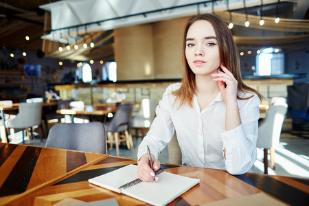 Portrait of beautiful young businesswoman looking at camera while sitting at table in cafe and making notes in her notebookの写真素材
