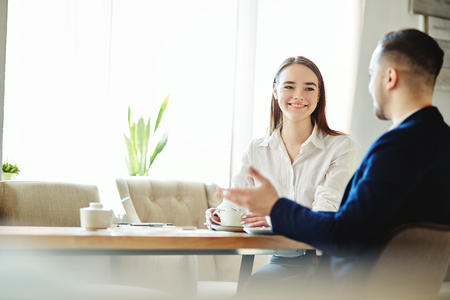Portrait of smiling young businesswoman listening to male coleague while sitting at table during coffe break in cafeの写真素材