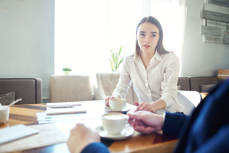 Young businesswoman talking to unrecognizable male colleague during coffee meting in cafe, documents and notebooks on tableの写真素材
