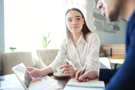 Young businesswoman and her unrecognizable male collague discussing data on tablet computer screen during meeting at cafe. Woman holding tablet and man making notes in his notebookの写真素材