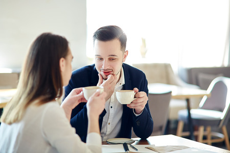 Two business people, young man and woman, sitting at table in cafe and having coffee. Businessman answering question from female colleagueの写真素材