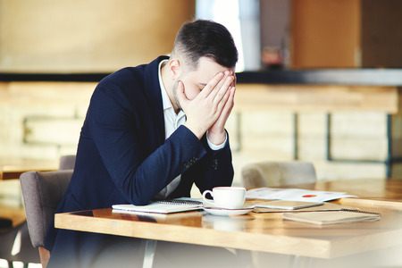 Frustrated young businessman in suit sitting at table in cafe with his hands over face, feeling stressed and exhaustedの写真素材