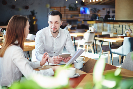 Business partners planning project at cafe. Businesswoman with documents in her hands and businessman with tablet computer sitting at table and talkingの写真素材