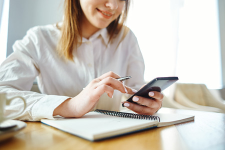 Young businesswoman working at table in cafe. Female manager holding smartphone over open notebook and using mobile applicationの写真素材