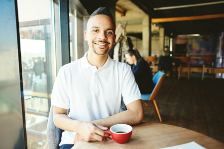 Portrait of cheerful mixed race man smiling at camera while having coffee at cafe tableの写真素材