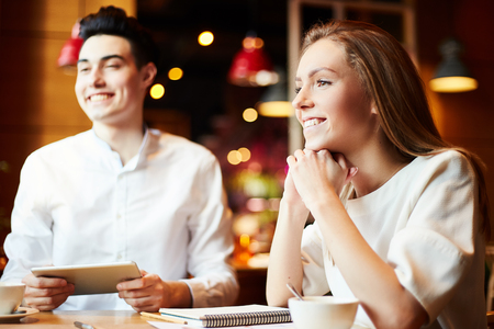 Young cheerful man and woman sitting with notepads and coffee cups at table in cafe, looking away and smilingの写真素材