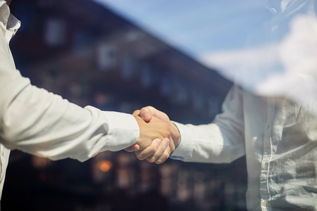 Shot through reflective glass of crop businessmen shaking hands in agreement standing in sunshineの写真素材