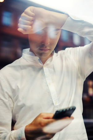 View through shiny glass of modern businessman in white shirt leaning on window and surfing phoneの写真素材