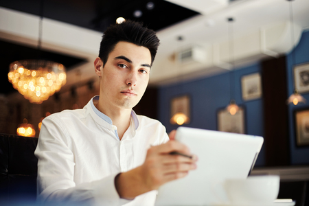 From below of handsome serious man in formal shirt sitting at table with tablet in cafe looking at cameraの写真素材