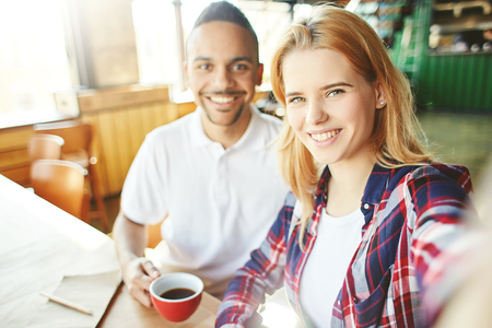 Friendly fellow stundets or young coworkers, Caucasian woman and Latin American man, taking selfie together at coffee breakの写真素材