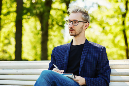 Blond male with creative haircut in glasses and dark checkered jacket sitting with notebook and pen on white park bench and looking away on blurred background at summerの写真素材