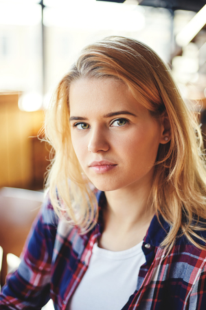 Headshot portrait of serene young woman with blond hair looking at camera. Girl wearing white t-shirt and checkered shirtの写真素材