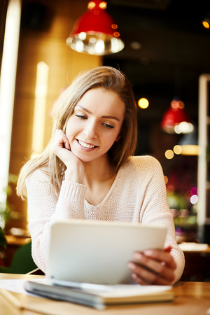 Beautiful young woman sitting at table in cafe and looking at tablet screen with smile, her hand on chinの写真素材