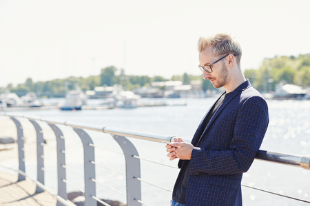 Relaxed young handsome man in glasses and dark jacket standing with mobile phone and leaning back on shiny metal handrail on sunny day with water and boats on blurred backgroundの写真素材