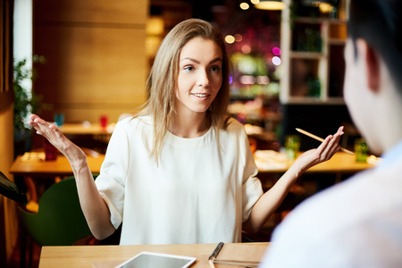 Young woman holding pencil and explaining something expressively while talking to man at table in cafeの写真素材