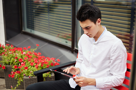 Modern young businessman in white shirt sitting on bench on terrace with flowers and surfing tabletの写真素材