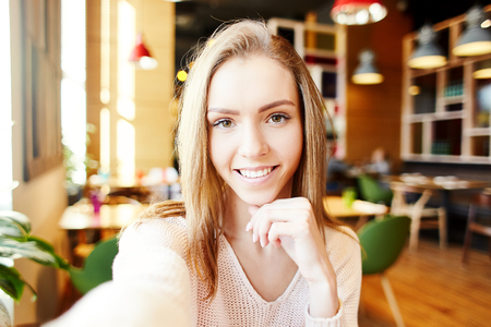 Young woman smiling at camera while taking selfie on background of cafe interior, her hand on chinの写真素材