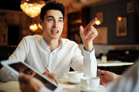 Young man sitting at table with anonymous coworker having business meeting with coffeeの写真素材